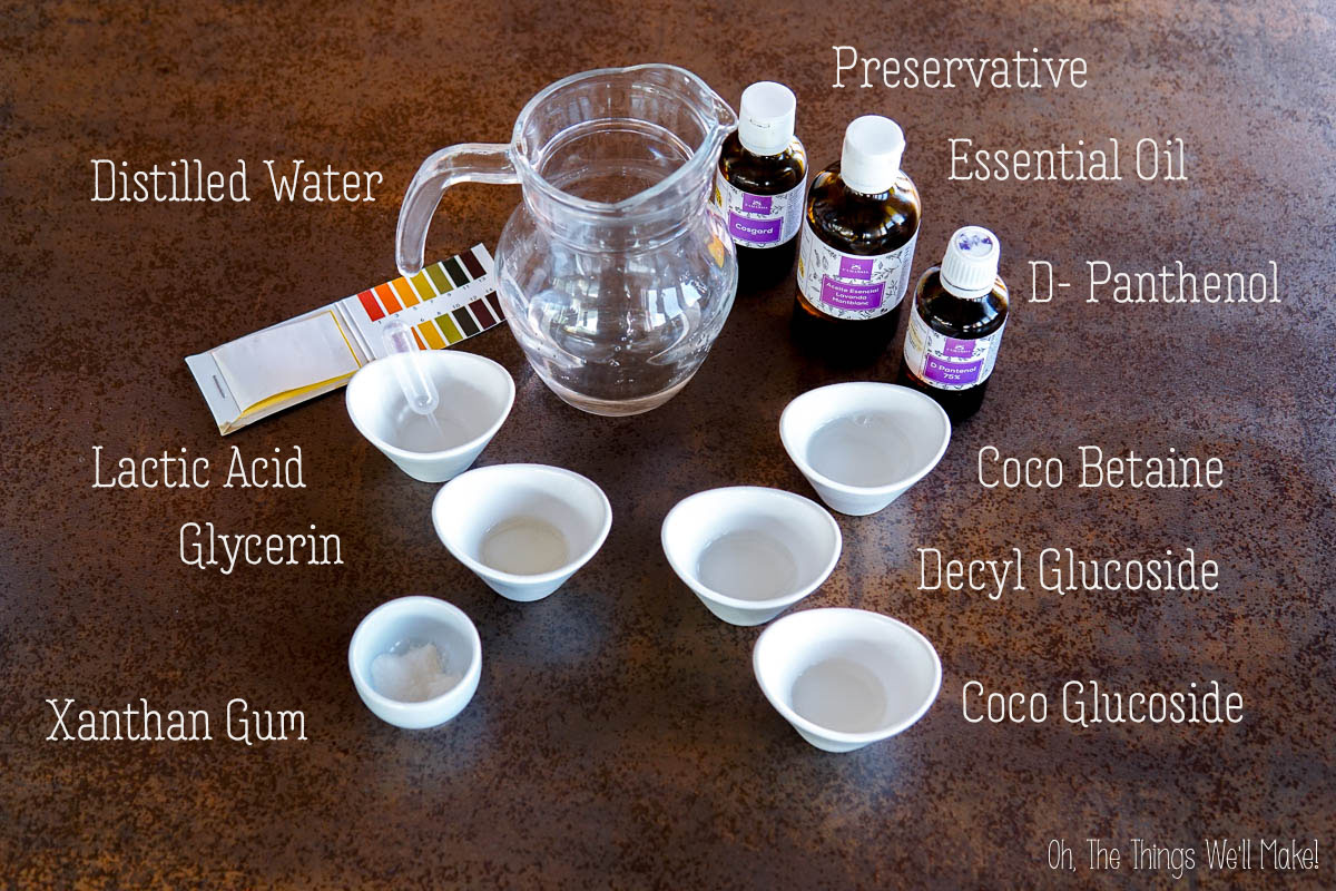 A pitcher of distilled water, bottles, labeled bowls of ingredients, and pH test strips arranged on a tabletop for a DIY daily use shampoo cosmetic formulation.